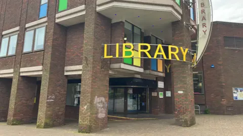 The outside of Redditch Library - showing five brick pillars and a main entrance, with a large yellow sign above it that says 'library'.