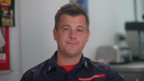 Jamie Niblock/BBC Adam Willis sitting down in a room with white walls. He is looking directly at the camera and smiling and is dressed in fire service uniform.