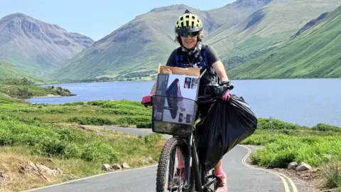 Lindsay Buck looks into the camera and smiles as she rides her bike up a hill. She is wearing a yellow helmet and black T-shirt. A basket on the front of her bike is filled with cardboard and a full black refuse sack is hanging from the handle. A lake and mountains can be seen in the background.