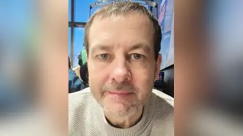 A man wearing a grey knit sweater stairs into the camera. He has short brown hair and grey stubble on his upper lip and lower face. Behind him is a clear window, a blue sky can be seen outside. 