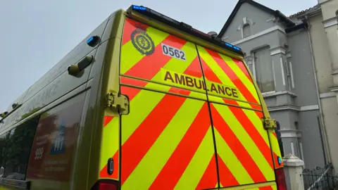 Rear view of a parked ambulance with bold yellow and red diagonal stripes. The word 'AMBULANCE' is prominently displayed in black capital letters across the back doors. Above it, the number '0562' and an emblem are visible. Residential buildings are in the background.