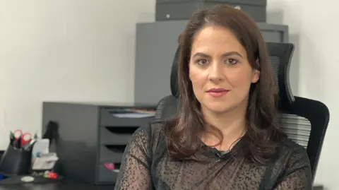 Claudia is seated at an office desk in a swivel chair, facing the camera. She is wearing a black, semi-sheer long-sleeved top. Behind the desk are office items including document trays, a printer, stationery holders, and storage cabinets against a light-colored wall.
