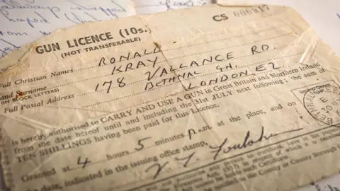 Bearnes Hampton & Littlewood A piece of paper with the words gun licence at the top of it. The name Ronald Kray are written in by hand and his address is given as 178 Vallance Road, Bethnal Green, London, E2.
It also has an official date stamp of 30 May visible to the right and the signature of the official who issued the licence.
It was issued at '4 hours 5 minutes pm'.