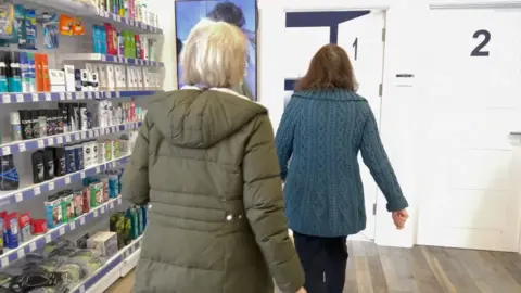 NICOLA HASELER/BBC Dr Karen Benson calls a patient through to her consulting room within the pharmacy. To the left you can see a shelf full of deodorant and shampoo within the pharmacy. The doctor walks towards the door marked number 1 in the centre of the picture with her back to the picture. The patient is following her wearing a khaki green puffer coat. The patient has grey hair in a bob.