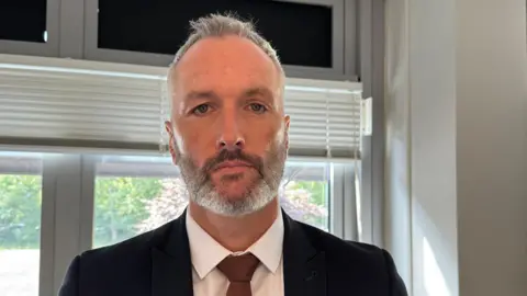 Det Con Lee Harshey-Jones. He looks at the camera with a neutral expression, and wears a white shirt, black suit jacket and bronze tie. He has short grey hair and short stubble. 