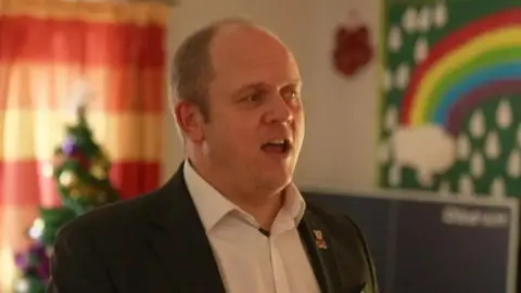 BBC Gareth Pierce looks away from the camera and speaks. He wears a white shirt and a dark jacket with a teddy bear badge. A Christmas tree and a colorful rainbow mural can be seen in the background of a room in the hospice.