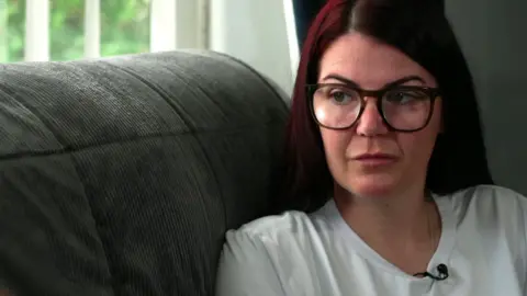 Carly Calland with long dark auburn hair and black glasses wearing a white t-shirt sitting on a grey couch in front of a window looking to the side.