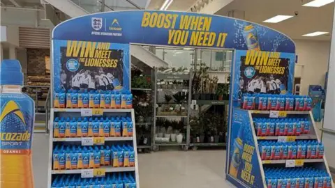 Jamie Davies A blue stand in a supermarket selling bottles of Lucozade sport. It has two sides and an archway, which reads 'boost when you need it' in yellow writing. Either side it has a promotion saying 'Win and meet the Lionesses' with a picture of the England women's team