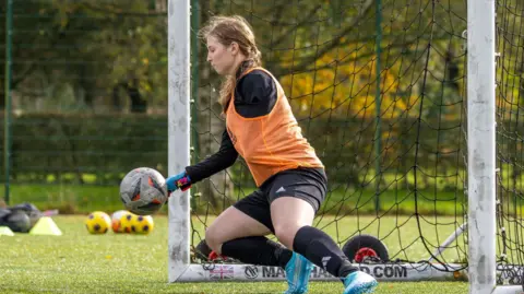 PDR Photos A goal keeper on the team, who has an amputated arm, catches a ball with her hand. She is bending down to catch the ball while in goal. She wears an orange vest over a black t-shirt, as well as black shorts, black socks, and blue trainers.