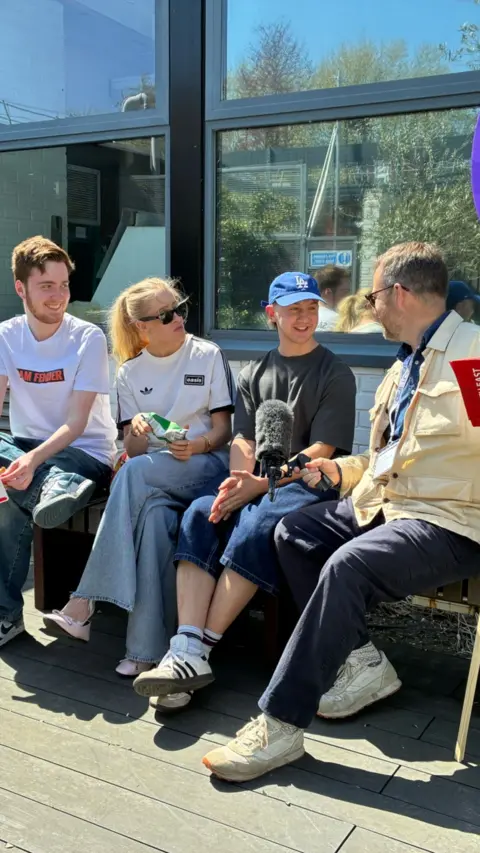 Four people sat in a bench in London. One is holding a michrophone.