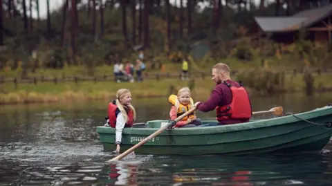 Center Parcs Two children in a boat with a man, all wearing lifejackets, on a pond with a grassy bank in the background