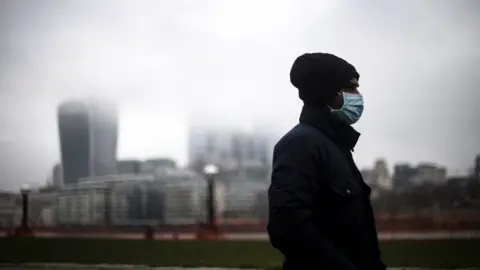 PA Media Man walks along south bank of Thames through city of London on a grey day, with a mask on