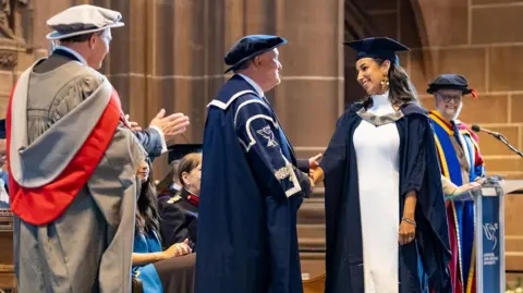 Fotografía de la Universidad John Moores de Liverpool. Charnele Washbourne cruza el escenario en su ceremonia de graduación luciendo un vestido de novia, con su toga y birrete, dentro de la Catedral Anglicana de Liverpool. Saluda con un apretón de manos a los funcionarios de la Universidad John Moores de Liverpool. Sonríe, al igual que los funcionarios presentes en el escenario.