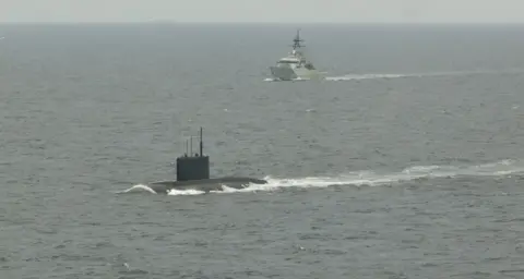 A submarine at the front of the image in the water with HMS Mersey in the distance