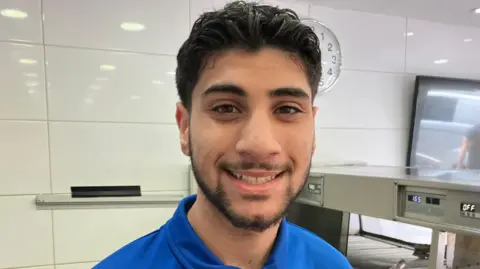A young man with dark hair, skin and eyes wearing a blue t-shirt and standing in front of a metal fish-and-chip shop fryer smiling.