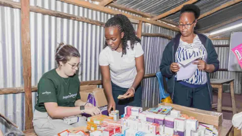 Ella McLaughlin is sitting at a wooden table covered with dozens of small boxes of medication. Two Kenyan women are standing alongside her, helping to organise things.