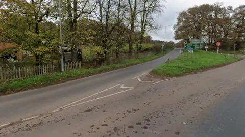 Google A Google street view image of a junction in the countryside. A road breaks off to the left past some trees and a wooden fence. Road signs can be seen on a triangular grass island between three roads.