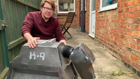 James Grant/BBC Man with purple shirt and glasses poses next to metallic dog