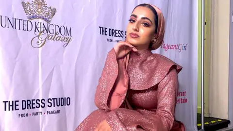 Fatimah Bobrah  Fatima is wearing an glamourous pink long sleeved dress and matching hijab. She has a nose ringe piercing and has her right hand under her chin