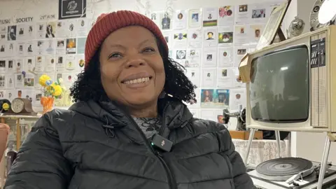 Jon Wright/BBC A woman in a hat sits in front of an exhibition of local Windrush generation people who have passed away.