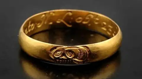 Hansons Auctioneers A gold ring resembling a wedding band reflecting on a polished black surface. It has a skull engraved on the top and an ornate inscription on the inside.