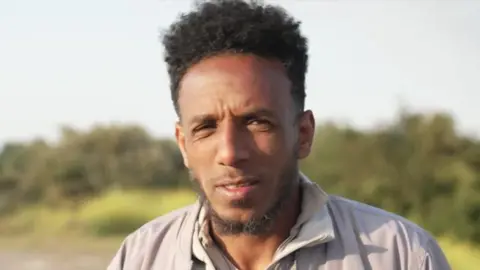 A man with dark hair and a thin dark beard. He is wearing a grey coat and there are green bushes in the background.