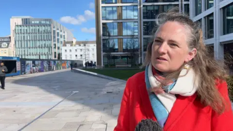 BBC Deputy Hilary Jeune is wearing a red coat with a blue and white scarf. She is stood in Trenton square in Jersey's finance centre. 