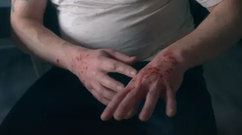 John Bell, pictured from the chest down, shows his hands with cuts and blood on them. He is wearing a white T-shirt and black pants. 