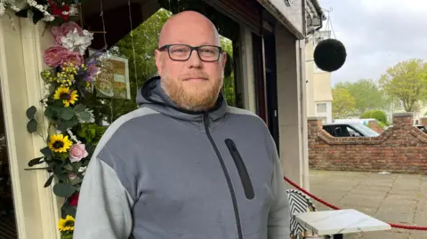 James stands for the photo outside his coffee shop, near to the proposed plans for new cycle infrastructure. He is wearing a grey hoodie, is bald and has black glasses.
