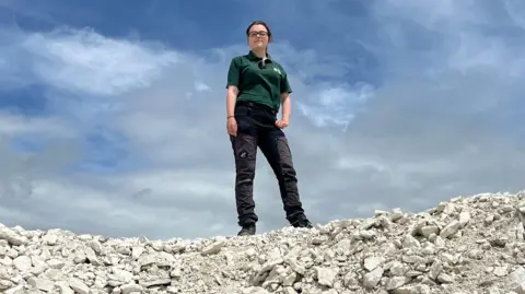 A woman in black trousers and green polo shirt stands on top of a pile of chalk looking at the camera on a sunny day. The sky is visible behind her.