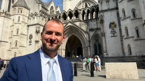 Tim Bearder has short brown hair and a brown beard. He is wearing a blue blazer and jumper over a pale blue tie and white shirt.