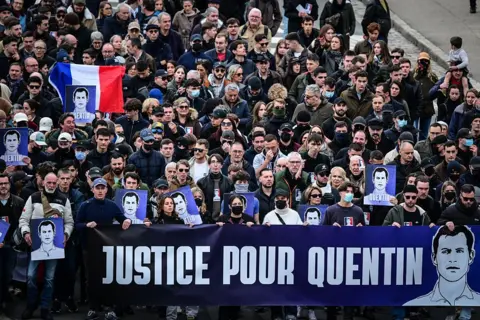 Getty Images A large crowd march behind a banner which reads "Justice pour Quentin" with an sketch portrait of him. One protestor carries the French flag. 