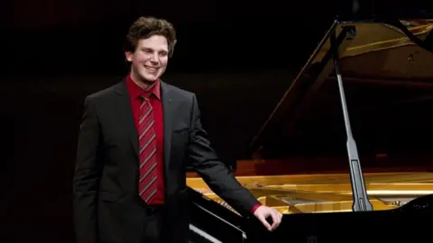 British-Australian pianist Jayson Gillham wearing a suit stands beside a grand piano.