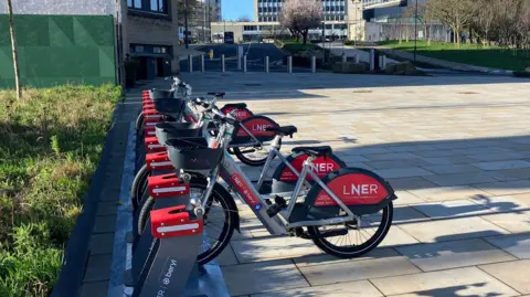 Four bikes and about 10 bike docks in grey and red. They are in a pedestrianised street area with grass to the left and pale paving stones to the right