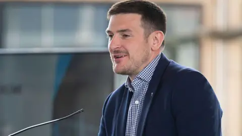 James Evans is stood wearing a blue suit and blue checkered shirt which is undone at the top, and is looking off to the left where he is speaking into a small microphone.