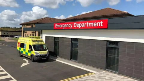 BBC A single-storey building with an ambulance parked outside. The building has a red sign that says "Emergency Department" on it.