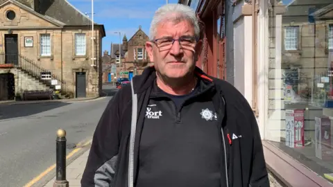 BBC A man in a black top and jacket stands outside a shop with a historic Tolbooth building in the background