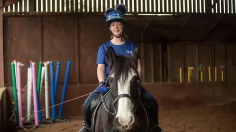 StoryShop Sheree sits astride a dark brown horse in a riding hall. She wears a blue and yellow tartan My Name's Doddie hat and a blue top, jeans and riding boots as well.