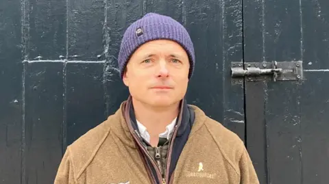 Johnny Wake look at the camera wearing a purple hat and brown fleece. He is in front of a large painted dark grey wooden door with a metal bolt to the right.