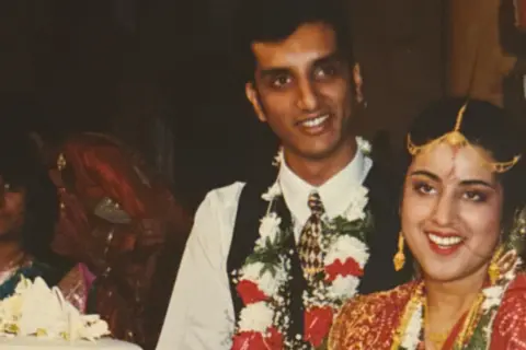 Sarju Patel Sarju and his wife are pictured on their wedding day, cutting a three tiered wedding cake. His wife is wearing an intricately designed Panetar, the traditional white and red Gujarati bridal saree, complemented by a Gharchola, a rich red silk saree with bandhni work often draped over the Panetar as part of Gujarati wedding customs. Sarju is dressed in a white shirt with a black waistcoat and tie.