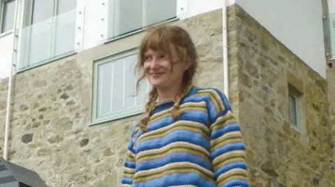 Other A woman with a blue striped jumper and braided hair stands next to a coastal building and smiles.