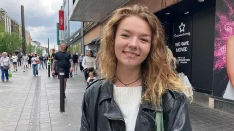 Head and shoulder shot of Tixie Wynn. She has long curly blonde to light brown hair, is smiling and has facial piercings on her eyebrow and chin. She is wearing a dark leather jacket over a white jumper. Behind her people can be seen walking on a busy shopping street, with shopfronts to the side of her.