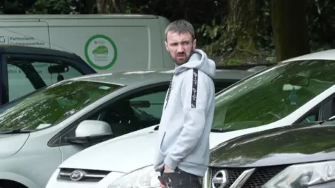 A man with brown hair and beard, wearing a grey hoodie. He is stood in front of cars and looking off to the side with a frown.