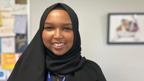 BBC News/Hayley Clarke Hafsa Yousef smiles at the camera, infront of a school noticeboard