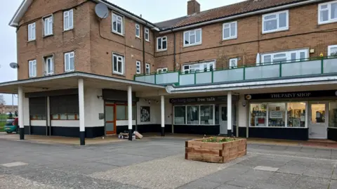 West Northamptonshire Council A freshly painted Park Square, with a wooden planter in the middle. There are several shops and flats above the building with a glass barrier in front of the flats. A car is parked to the left with a play area behind the car. A satellite dish is on a wall and there are lots of white windows to the flats. 