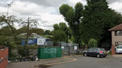General view of Ward End Park near the Foxton Road entrance, It shows a green street cabinet mental fences and a gathering of trees. A grey car is visible in the foreground