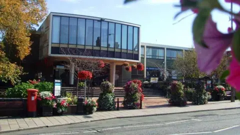 Staffordshire Police A municipal building on a street in a town, with manicured bushes and a red postbox outside. The building is two storey, with a large glass frontage on the second floor, which is supported by pillars. Steps lead up to a receded entrance.