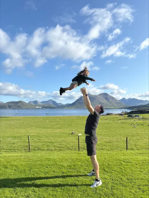Laura Millar A man in dark shorts and T-shirt standing in a grassy field with a loch and hills in the distance, throwing a small toddler-aged girl in the air