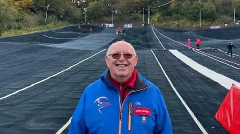 John Lowther stands in front of Kendal Snowsports club which is a dry slope. He is wearing sun-tinted glasses, a branded blue coat and a red fleece. He has short silver hair. There are people in the background being taught how to ski.