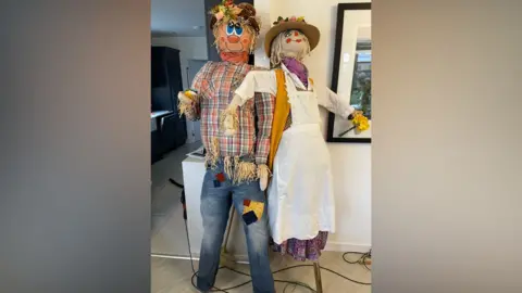 Chatteris in Bloom Two scarecrows are leaning against a wall inside of house. The scarecrow on the left is wearing a tartan shirt and blue jeans which have patches on them. The scarecrow on the right is wearing a purple dress with a white apron over the top.